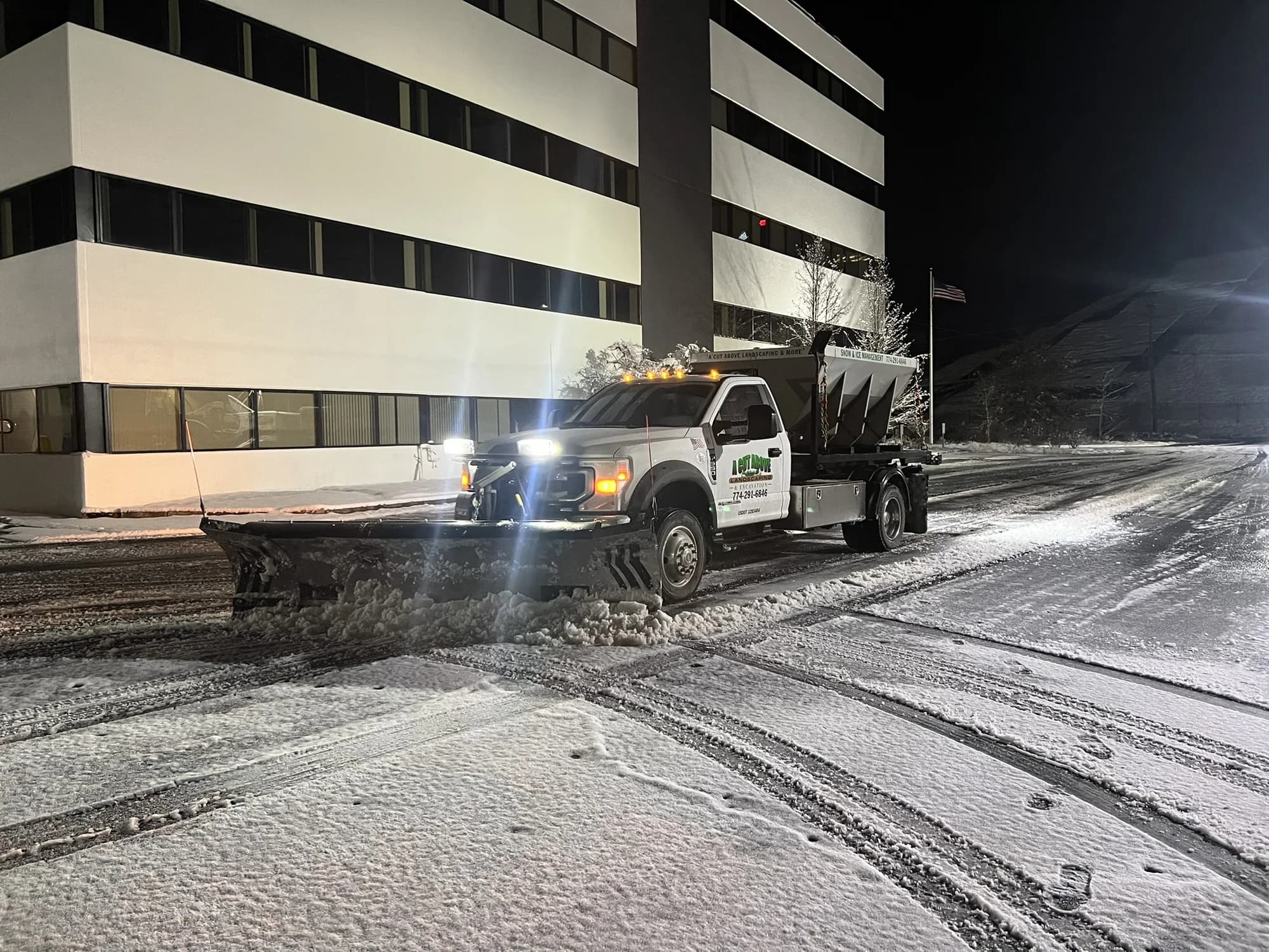 A Cut Above plow truck clearing a snowy access road beside a parking garage at night
