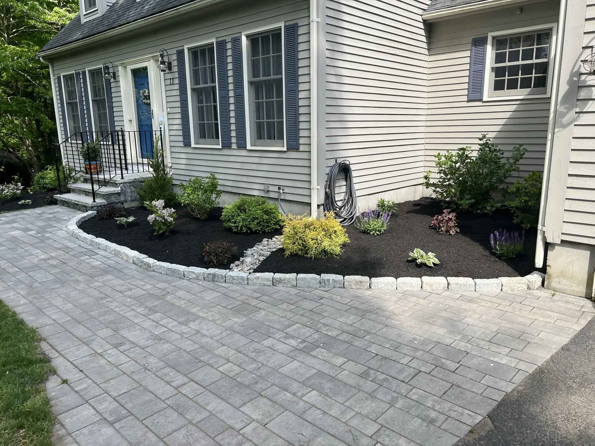 Wide gray paver entrance beside cobblestone-edged planting beds with shrubs and flowers