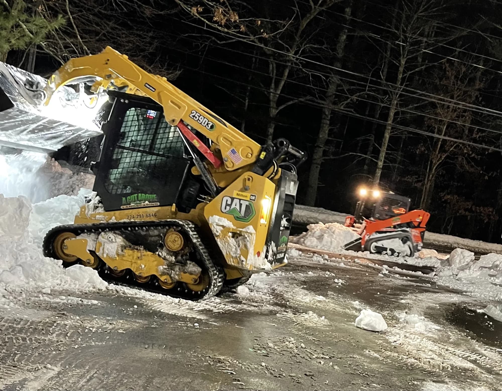 Two compact tracked loaders stacking snow beside a cleared pavement area at night