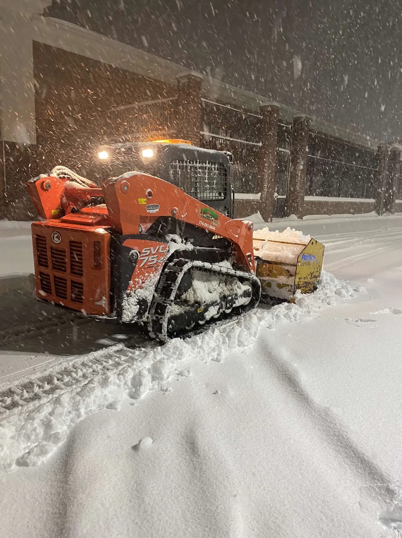 Compact tracked loader pushing snow beside a commercial building during nighttime snowfall