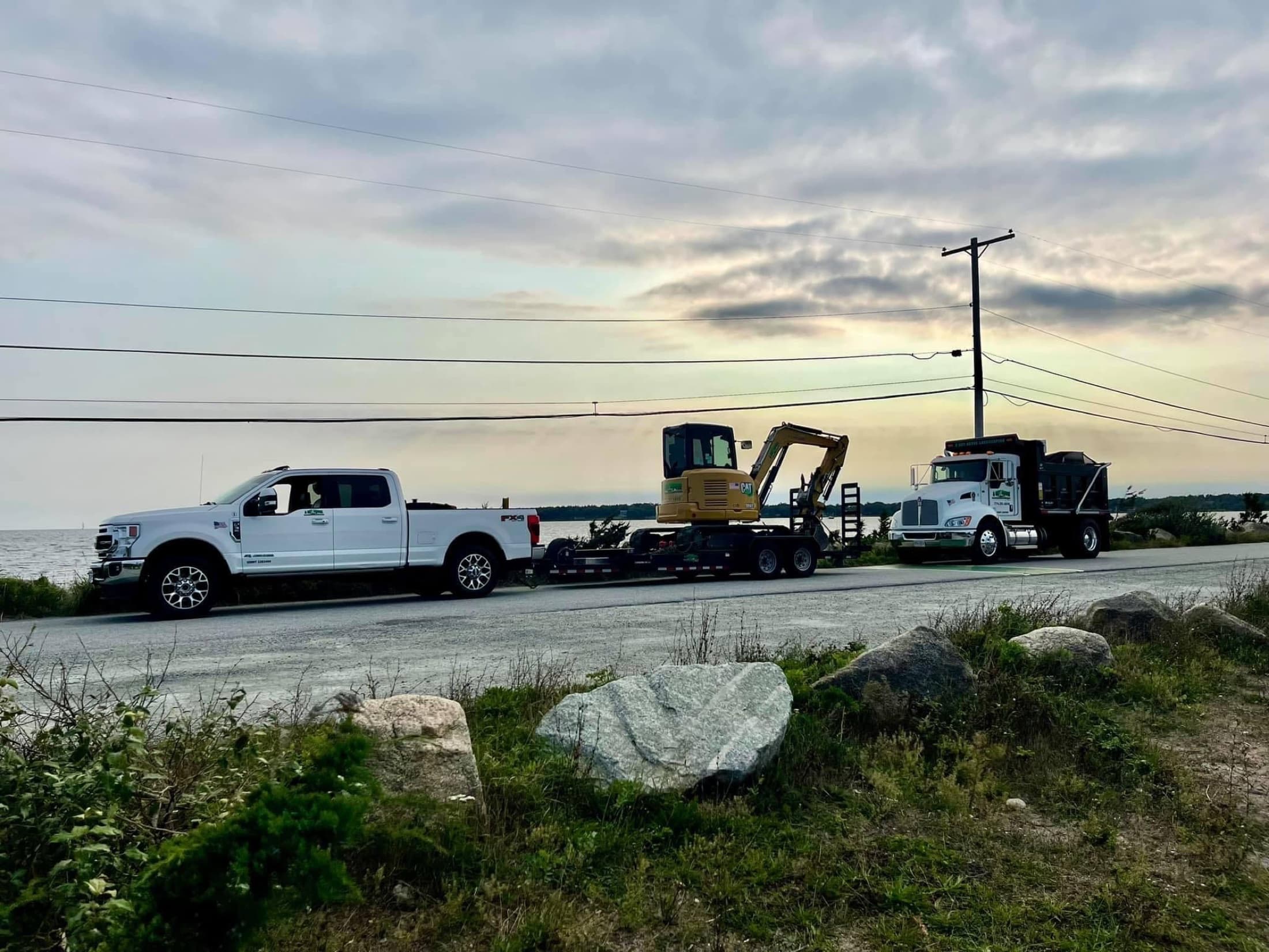 A Cut Above work trucks and excavator traveling together beside the water